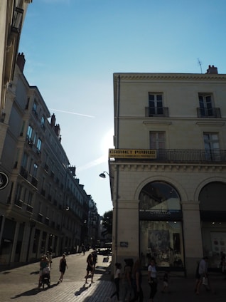 A sunlit street with people walking along a cobblestone path. On the right, there is a building with a sign that reads 'Cabinet Piroud' and has arched windows. The sky is clear with a visible contrail and the sunlight creates strong contrasts and shadows.