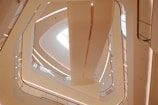 A modern architectural interior featuring an upward view of several floors with clean, curved lines and geometric shapes. The design includes multiple balconies or walkways lined with glass and metal railings, illuminated by both natural and artificial lighting.