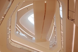 A modern architectural interior featuring an upward view of several floors with clean, curved lines and geometric shapes. The design includes multiple balconies or walkways lined with glass and metal railings, illuminated by both natural and artificial lighting.