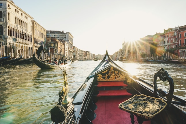 A traditional gondola navigating a sunlit canal lined with historic buildings. The sun is setting behind, casting a warm glow over the water and architecture.