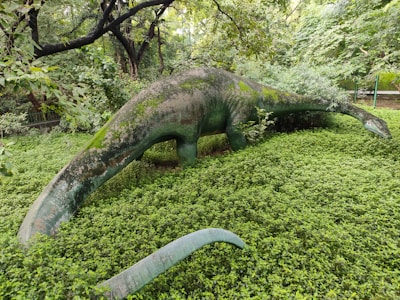 A large dinosaur sculpture stands among dense greenery, partially covered by moss and surrounded by lush plants. Overarching trees frame the scene, creating a natural and wild setting.