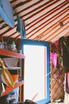 A cozy interior of a yurt with a wooden lattice structure and a blue door partially open to the outside. Shelves are lined with various kitchen items including bowls, a strainer, and pitchers. Clothing hangs along the wooden poles, and colorful textiles add warmth to the space.