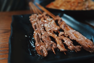 Skewered pieces of grilled meat, likely beef or lamb, are arranged on a black rectangular plate. The meat is lightly garnished with sesame seeds and appears juicy and well-cooked. The background includes a blurred view of more food in a round dish.