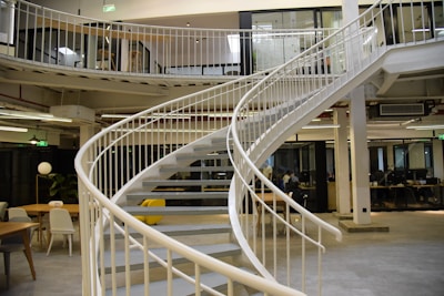 A modern office space features a spiral staircase with white railings, leading to an upper floor. The space is open, with several tables and chairs, and a few people working at their desks. The design incorporates industrial elements like exposed pipes and large windows.