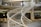 A modern office space features a spiral staircase with white railings, leading to an upper floor. The space is open, with several tables and chairs, and a few people working at their desks. The design incorporates industrial elements like exposed pipes and large windows.