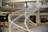 A modern office space features a spiral staircase with white railings, leading to an upper floor. The space is open, with several tables and chairs, and a few people working at their desks. The design incorporates industrial elements like exposed pipes and large windows.