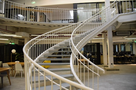 A modern office space features a spiral staircase with white railings, leading to an upper floor. The space is open, with several tables and chairs, and a few people working at their desks. The design incorporates industrial elements like exposed pipes and large windows.