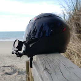 A black motorcycle helmet with a mounted camera is resting on a wooden railing. In the background, there is a sandy beach with gentle waves and some beach grass on the side.