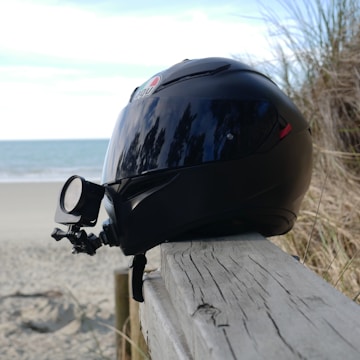 A black motorcycle helmet with a mounted camera is resting on a wooden railing. In the background, there is a sandy beach with gentle waves and some beach grass on the side.