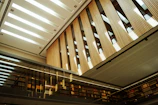 A modern interior space featuring tall wooden panels and a sleek, glass façade. The ceiling is designed with rectangular cutouts allowing natural light to illuminate the space. Bookshelves line the walls, and the reflective surfaces create an open and airy atmosphere.