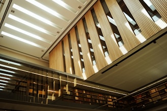 A modern interior space featuring tall wooden panels and a sleek, glass façade. The ceiling is designed with rectangular cutouts allowing natural light to illuminate the space. Bookshelves line the walls, and the reflective surfaces create an open and airy atmosphere.
