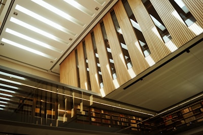 A modern interior space featuring tall wooden panels and a sleek, glass façade. The ceiling is designed with rectangular cutouts allowing natural light to illuminate the space. Bookshelves line the walls, and the reflective surfaces create an open and airy atmosphere.