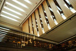 A modern interior space featuring tall wooden panels and a sleek, glass façade. The ceiling is designed with rectangular cutouts allowing natural light to illuminate the space. Bookshelves line the walls, and the reflective surfaces create an open and airy atmosphere.