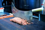 A large meat cleaver with a white handle is embedded in a block of minced meat on a dark wooden cutting board. The background reveals a kitchen setting with blurred stainless steel equipment and a container of vegetables.