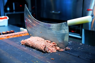 A large meat cleaver with a white handle is embedded in a block of minced meat on a dark wooden cutting board. The background reveals a kitchen setting with blurred stainless steel equipment and a container of vegetables.