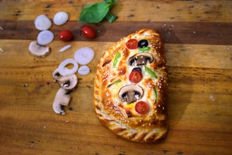 A golden-brown calzone with a braided crust sits on a wooden board. It is topped with sesame seeds, slices of mushrooms, cherry tomatoes, black olives, and green bell peppers. To the left, there are sliced onions, mushrooms, and whole cherry tomatoes, along with basil leaves.