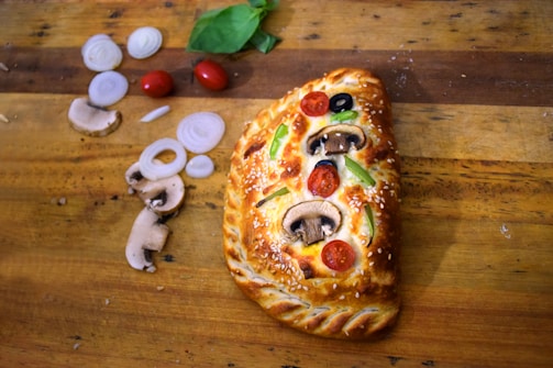 A golden-brown calzone with a braided crust sits on a wooden board. It is topped with sesame seeds, slices of mushrooms, cherry tomatoes, black olives, and green bell peppers. To the left, there are sliced onions, mushrooms, and whole cherry tomatoes, along with basil leaves.