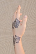 An intricately tattooed hand is shown, with black geometric and swirling patterns on the wrist and back of the hand, set against a beige, textured circular background.