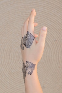 An intricately tattooed hand is shown, with black geometric and swirling patterns on the wrist and back of the hand, set against a beige, textured circular background.