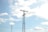 A tall metal transmission tower is set against a backdrop of a clear blue sky filled with scattered white clouds. Power lines extend horizontally across the image connecting to the tower, indicating its function in electricity distribution.