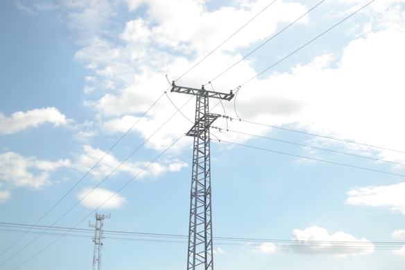 A tall metal transmission tower is set against a backdrop of a clear blue sky filled with scattered white clouds. Power lines extend horizontally across the image connecting to the tower, indicating its function in electricity distribution.