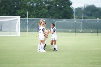 A group of female athletes, dressed in white uniforms, are standing on a soccer field. They are participating in some sort of team ritual or conversation, with one of the players holding a bat-like object. The background features a goal post and a chain-link fence with green coverings, while trees are visible in the distance.