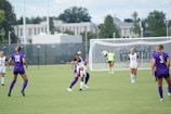 A women's soccer match is taking place on a grassy field with players from two teams, one in purple and the other in white uniforms. The focus is on two players challenging for the ball mid-air, while other players observe or prepare for the next move. In the background, a goalkeeper stands near the goalpost, and part of the slogan 'DON'T GIVE UP THE SHIP' is visible on a banner. Trees and buildings are visible beyond the fence surrounding the field.