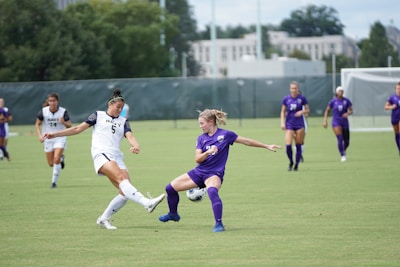 Two soccer players contest the ball on a lush green field, surrounded by a few other players in the distance. The player in white is mid-kick, while the player in purple engages defensively, with focused determination on their faces. The scene is set in an outdoor stadium with a backdrop of trees and buildings under a partly cloudy sky.