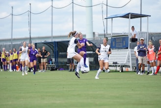 Soccer players are engaged in a match on a grassy field. One player is jumping to attempt a kick, while others are running nearby. Players in white and blue uniforms are visible. To the side, there are coaches and substitutes observing the game.