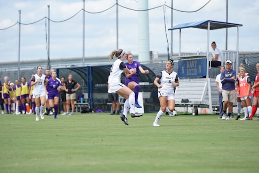 Soccer players are engaged in a match on a grassy field. One player is jumping to attempt a kick, while others are running nearby. Players in white and blue uniforms are visible. To the side, there are coaches and substitutes observing the game.