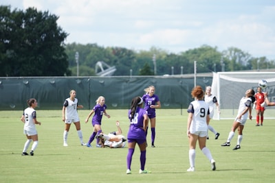 A group of female soccer players are actively engaged in a match on a grass field. Some players are wearing white uniforms with navy details, while others are in purple. One player is heading the ball in mid-air with others watching or running in the background. A goalie dressed in red is positioned near the goalpost. A large net, trees, and a cloudy sky serve as the backdrop.