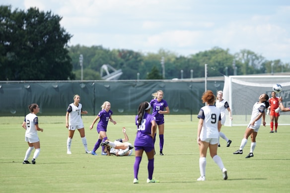 A group of female soccer players are actively engaged in a match on a grass field. Some players are wearing white uniforms with navy details, while others are in purple. One player is heading the ball in mid-air with others watching or running in the background. A goalie dressed in red is positioned near the goalpost. A large net, trees, and a cloudy sky serve as the backdrop.