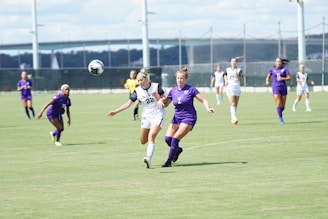 Two soccer players, one in a white uniform and the other in a purple uniform, are running on a grass field while competing for a soccer ball in mid-air. Other players wearing similar uniforms can be seen in the background, along with a referee in a yellow shirt. A large structure, possibly a bridge, is visible in the distance under a partly cloudy sky.