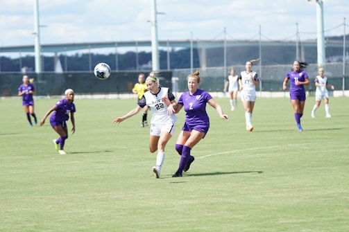 Two soccer players, one in a white uniform and the other in a purple uniform, are running on a grass field while competing for a soccer ball in mid-air. Other players wearing similar uniforms can be seen in the background, along with a referee in a yellow shirt. A large structure, possibly a bridge, is visible in the distance under a partly cloudy sky.