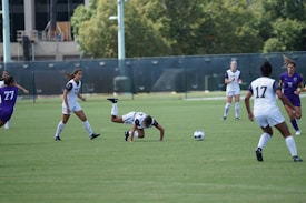 A group of female soccer players on a grassy field, actively engaged in a match. Several players are wearing white uniforms with some in purple. One player is in the middle of a mid-air fall at the center of the action, while others are focusing on the ball.