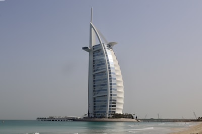 A modern, sail-shaped skyscraper stands prominently near the shoreline, surrounded by calm sea water and a clear blue sky. The structure is a notable architectural landmark with a unique design, situated on a man-made island connected by a bridge.