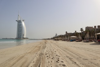 A vast sandy beach extends under a clear sky, with footprints marking the sand. To the left stands a prominent sail-shaped building near the water. On the right, rows of lounge chairs and umbrellas line the beach, with palm trees and a few modern buildings in the background.