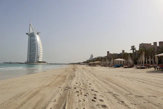 A vast sandy beach extends under a clear sky, with footprints marking the sand. To the left stands a prominent sail-shaped building near the water. On the right, rows of lounge chairs and umbrellas line the beach, with palm trees and a few modern buildings in the background.