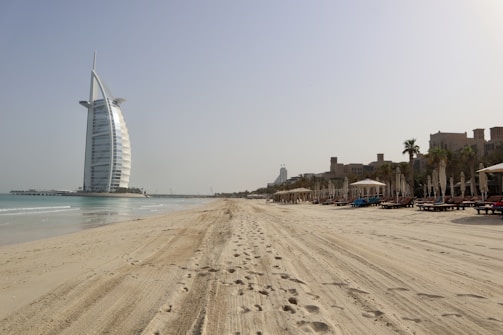 A vast sandy beach extends under a clear sky, with footprints marking the sand. To the left stands a prominent sail-shaped building near the water. On the right, rows of lounge chairs and umbrellas line the beach, with palm trees and a few modern buildings in the background.