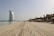 A vast sandy beach extends under a clear sky, with footprints marking the sand. To the left stands a prominent sail-shaped building near the water. On the right, rows of lounge chairs and umbrellas line the beach, with palm trees and a few modern buildings in the background.
