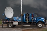 A large blue weather truck equipped with a radar dish and other scientific instruments is parked on a gravel road. Two people are standing on the side, one looking towards the sky and the other holding a clipboard inside the truck, with the doors open. Dark stormy clouds are visible in the background, suggesting severe weather conditions.