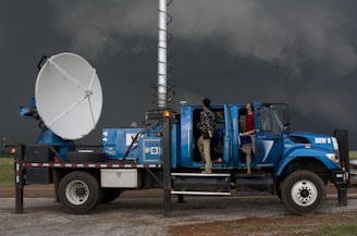 A large blue weather truck equipped with a radar dish and other scientific instruments is parked on a gravel road. Two people are standing on the side, one looking towards the sky and the other holding a clipboard inside the truck, with the doors open. Dark stormy clouds are visible in the background, suggesting severe weather conditions.
