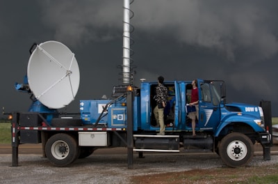 A large blue weather truck equipped with a radar dish and other scientific instruments is parked on a gravel road. Two people are standing on the side, one looking towards the sky and the other holding a clipboard inside the truck, with the doors open. Dark stormy clouds are visible in the background, suggesting severe weather conditions.