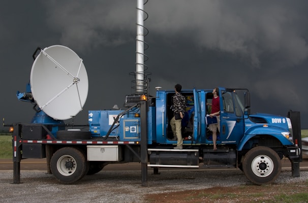 A large blue weather truck equipped with a radar dish and other scientific instruments is parked on a gravel road. Two people are standing on the side, one looking towards the sky and the other holding a clipboard inside the truck, with the doors open. Dark stormy clouds are visible in the background, suggesting severe weather conditions.