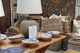A rustic interior setting features a collection of woven baskets stacked neatly in the background, accompanied by a selection of decorative ceramic bowls placed on a wooden table. A large, textured table lamp with a beige shade casts light over the scene, contributing to the warm ambiance. Artwork and a patterned textile decorate the walls, enhancing the room's artisanal feel.