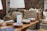 A rustic interior setting features a collection of woven baskets stacked neatly in the background, accompanied by a selection of decorative ceramic bowls placed on a wooden table. A large, textured table lamp with a beige shade casts light over the scene, contributing to the warm ambiance. Artwork and a patterned textile decorate the walls, enhancing the room's artisanal feel.