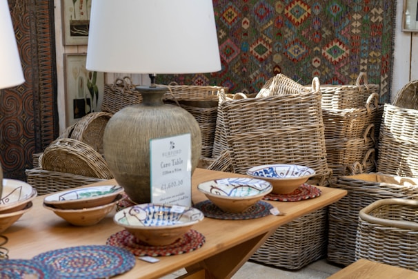 A rustic interior setting features a collection of woven baskets stacked neatly in the background, accompanied by a selection of decorative ceramic bowls placed on a wooden table. A large, textured table lamp with a beige shade casts light over the scene, contributing to the warm ambiance. Artwork and a patterned textile decorate the walls, enhancing the room's artisanal feel.