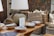A rustic interior setting features a collection of woven baskets stacked neatly in the background, accompanied by a selection of decorative ceramic bowls placed on a wooden table. A large, textured table lamp with a beige shade casts light over the scene, contributing to the warm ambiance. Artwork and a patterned textile decorate the walls, enhancing the room's artisanal feel.