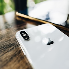 A close-up view of the back of a white iPhone with the Apple logo prominently displayed. The device is placed on a wooden surface, with soft, natural lighting creating reflections on the phone's surface. Another smartphone is visible in the blurred background, suggesting a casual setting.