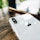 A close-up view of the back of a white iPhone with the Apple logo prominently displayed. The device is placed on a wooden surface, with soft, natural lighting creating reflections on the phone's surface. Another smartphone is visible in the blurred background, suggesting a casual setting.